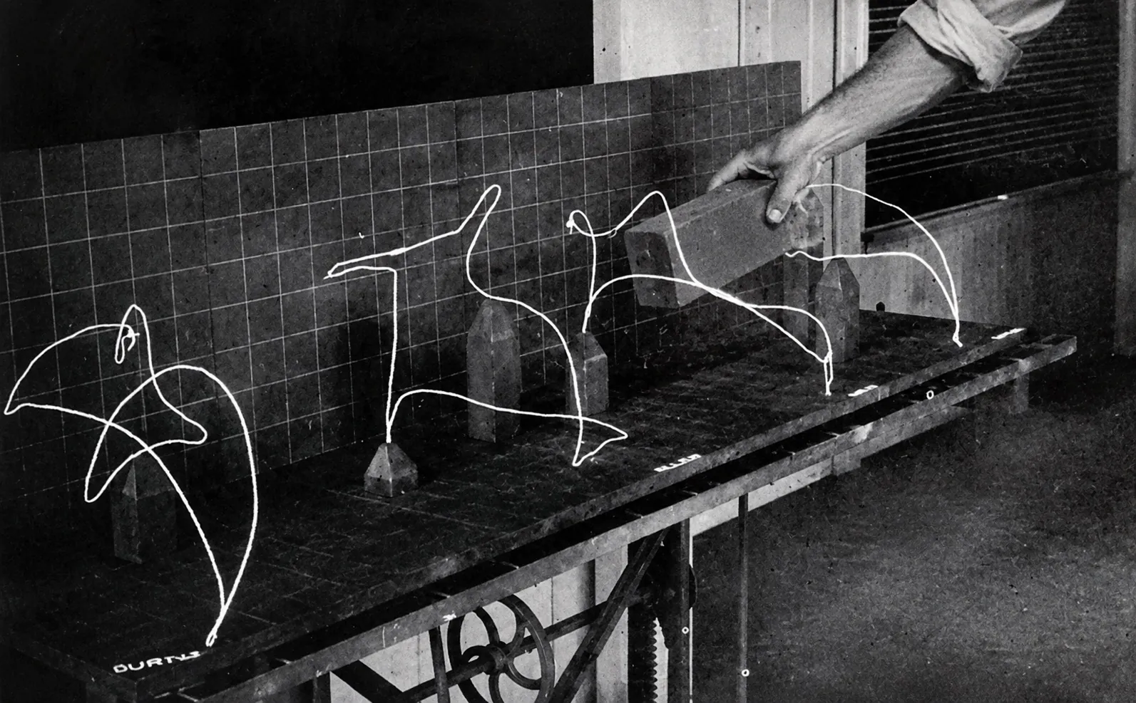 Wire models of a hand's path through the air,
                  standing in a row on a bench against a gridded backdrop, with a hand lowering a brick
                  past them.